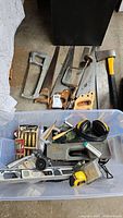Hand saws including Stanley, manual hand drill, axe with yellow and black handle, measuring tape, hearing protection earmuffs, paint brushes, Champion spark plugs, and hardware such as cabinet pull and clothing hooks photographed in garage.