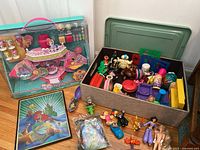 Wide shot of entire lot showing the My Little Pony boxed playset, fabric bin with assorted toys, small plastic figures, and a framed Ariel print on hardwood floor.
