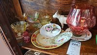 Shelf displaying various coloured glass stemware and vintage china pieces including teacup, plates, creamer, and cow-shaped creamer, along with a red glass goblet and a small red glass jar.