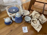 Overview of white porcelain tea set hand painted with blue peacock feather pattern and gold trim: teapot with lid, sugar bowl with lid, teacup and several saucers displayed on wrapping paper