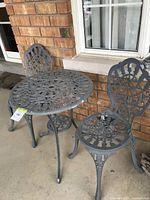 Three-piece gray cast aluminum bistro set against brick wall, shows round table and two chairs