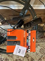 Top view of Amazon Fire TV Stick device, remote control, and power adapter with original orange box on glass table.