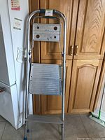 Foldable aluminum step ladder leaning against wooden kitchen cabinets, showing front with two wide steps and rounded handle.