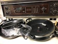 Five frying pans of various sizes; one frying pan has a glass lid with handle; all have black handles; set is placed on a stove top