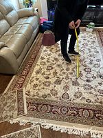 Rug laid on hardwood floor with person measuring width, showing ivory center, red and beige floral border, and white fringe.