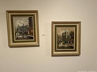 Two framed oil paintings hanging on a wall depicting urban scenes with people.