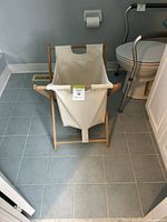 Laundry basket standing on a tiled bathroom floor next to a toilet, showing its wooden frame and linen sack with handle cutouts.