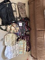 Overview of nine assorted Lesportsac bags and purses spread on floor next to brown sofa.