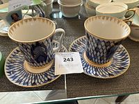 Two porcelain tea cups with matching saucers featuring cobalt blue and gold frieze patterns displayed on a shelf.