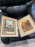 Two framed paintings placed on a black leather chair showing urban scenes with buildings and people, labeled with lot number 358.