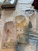 Three men's dress shirts unopened in plastic on a bed, showing packaging and brand labels