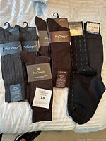 Seven pairs of men's dress socks arranged on a white surface. Five pairs by McGregor in brown and gray shades with labels attached. Two pairs black dress socks, one in a patterned style and one solid black by Star John.