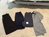 Four items of men's lounge clothing on a beige carpet: black pants, navy shorts with Puma logo, dark gray sweatpants, gray t-shirt with tag.