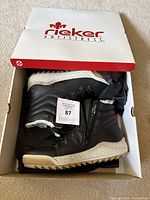 Pair of black Rieker snow boots inside branded shoebox, showing exterior side, sole, and part of the fur lining.