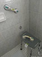 Photo of corner shower area showing two white suction cup hand grips mounted on the tiled wall. Gray adjustable shower stool with perforated seat placed below one of the grips.