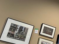Two framed art prints hung on a wall showing urban street scenes; the larger one is 20" x 16". Both prints are signed and numbered by the artist Dunant.
