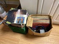 Photo of two stacks of vinyl records, one in a green crate and one in a worn cardboard box, showing records including ABBA's 'Super Trouper' and another with two men playing saxophones.
