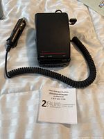 Top view of Cobra Trapshooter Radar Detector RD-2110 with coiled car charger cord attached, device placed on a fabric surface with an Alivio auction card below.