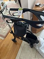 Side and top view of black walker showing ergonomic handles with hand brakes, mesh pouch, and front wheels on hardwood and tile floor background.