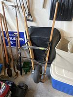 Wheelbarrow with black basin and single rubber wheel, wooden handles, indoors amongst other garden tools and storage tubs.