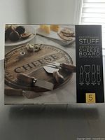 Photo of front of box showing the artisanal acacia wood cheese board with four cheese knives and 'CHEESE' engraving on board