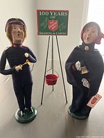 Two Byers' Choice Salvation Army caroller figurines, male and female, posed with metal donation bucket and sign in between on a flat surface.