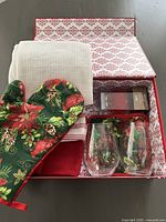 Overview of all items inside festive gift box: oven mitt, hot pad, tea towels, wine glasses, and bottle topper box.