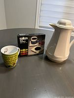 Salton electric mug warmer in box beside white ceramic coffee pot and green coffee mug.