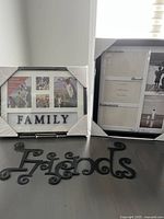 Two black collage picture frames wrapped in corner protectors, one displaying a view of the five photo openings and a 'FAMILY' label, placed upright on a flat surface against a wall. Below the frames lies a separate black 'Friends' sign made of durable plastic with decorative script.