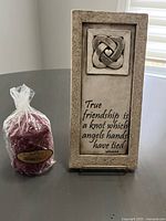 Photo of resin wall plaque with friendship knot design and inscribed quote, positioned beside wrapped red scented candle.