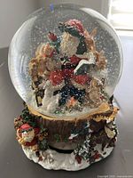 Full view of the waterglobe showing Santa inside the glass globe with animals and snow, and the decorated Christmas themed base resembling a wood stump with Santa figure and trees.