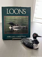 Front cover of the 'Loons' book with a color photograph of a loon with chicks and the loon-shaped coin bank in front.