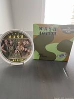 Photo of the M*A*S*H commemorative plate partially visible on stand next to its original box with themed camouflage design and labeling.