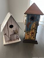 Photo showing two decorative birdhouses side by side: one white with a watering can image and metal roof, the other blue with a wooden door and crescent moon decoration