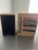 Front view of one speaker next to the original Paradigm Atom Oak box showing branding and model info
