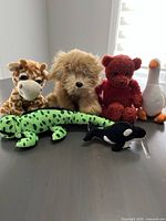 Photo showing 6 plush toys: giraffe, shaggy dog, red teddy bear, white goose, green spotted lizard and orca whale arranged together near window.