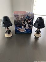 Pair of snowman tealight lamps with black metal shades and a box of snowman coaster set on a table.