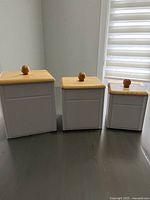 Three wood kitchen canisters of different sizes, white bodies, natural wood lids with round wooden knob handles, placed on dark surface near window with blinds.