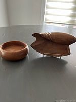 Two wooden serving dishes on a reflective surface under natural light: a round bowl and a shell-shaped tray with handle.