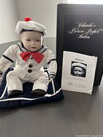 Full view of the Matthew porcelain doll sitting on a cushion alongside the Certificate of Authenticity and original box.