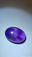 Oval cabochon deep dark purple amethyst gemstone under light, showing glossy polished surface and internal inclusions.