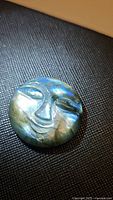 Close up image of a round labradorite stone carved into a smiling face with blue and green iridescent flash on dark textured background.