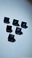 Six dark blue princess cut sapphires arranged in a loose group on a light surface under bright lighting revealing facets and clarity.