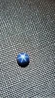 Close-up photo of a natural round blue star sapphire, showing the 6-ray star effect on its surface. The gemstone is set against a textured black background.