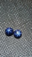 Pair of round blue star sapphire cabochon gemstones on black textured surface, showing distinctive six-ray star effect.