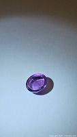 Oval cabochon cut natural Brazilian amethyst gemstone, purple color, about 10x8mm, shown on white background with clear light reflections.