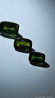 Three cushion-cut deep green chrome diopside gemstones arranged in a line on a light surface with visible facets and shine.