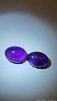 Two deep purple oval cabochon amethyst gemstones lying side by side on a light background, showing translucent color and some visible inclusions.