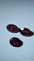 Three natural rhodolite garnet gemstones, faceted oval cut, showing pink-purple color with natural inclusions.