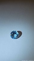 Close-up photo of a round brilliant cut natural sky blue topaz gemstone with vibrant light blue color on a plain background.